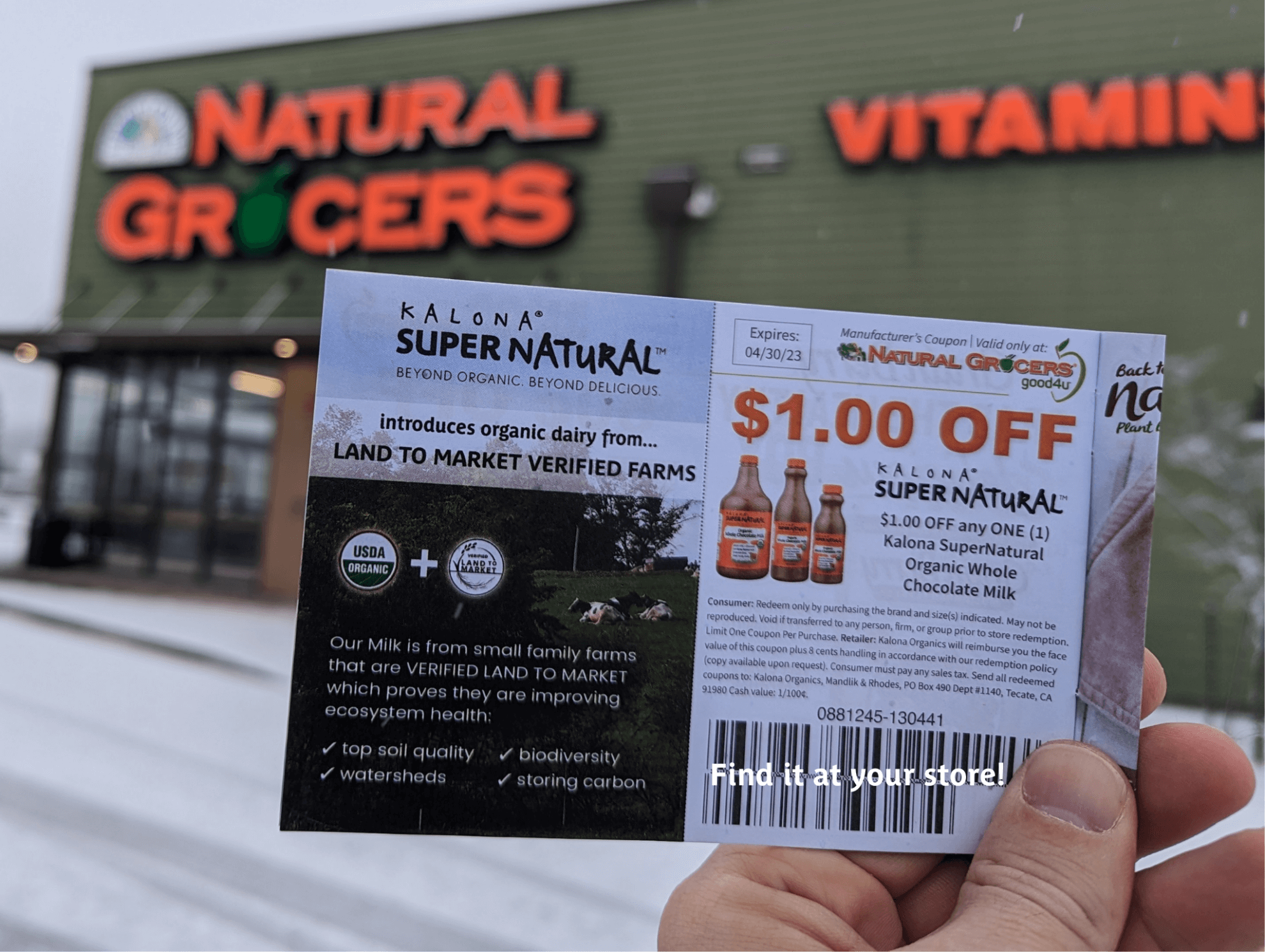 Kalona SuperNatural $1 Off Coupon held in front of Natural Grocers store