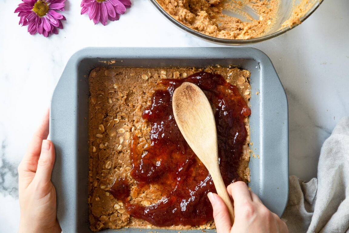 spreading jelly on bottom layer of peanut butter and oat mixture